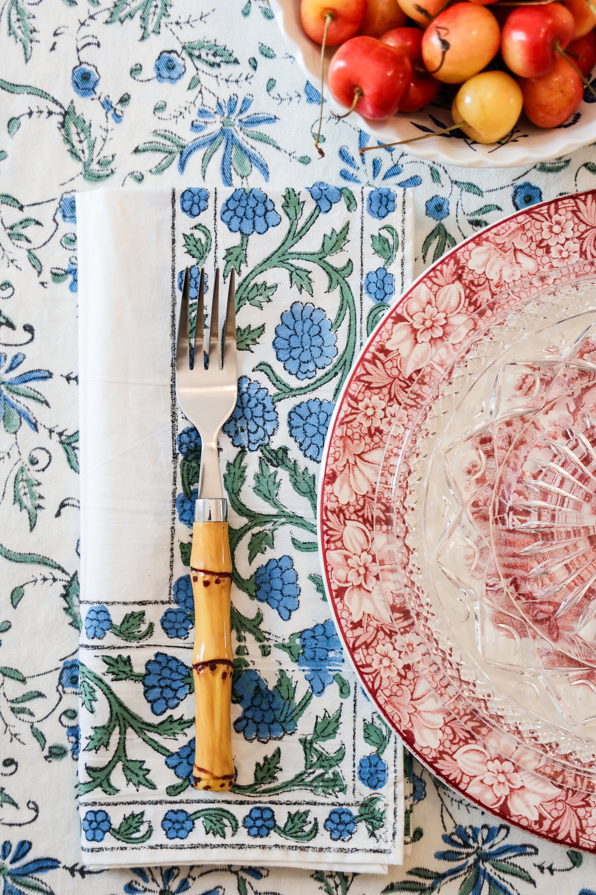 A set of dinner napkins with a floral pattern in blue and green, displayed on a table with a fork and knife, alongside a plate with red designs and an apple in the background.