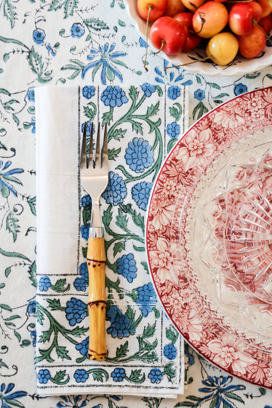 A set of dinner napkins with a floral pattern in blue and green, displayed on a table with a fork and knife, alongside a plate with red designs and an apple in the background.