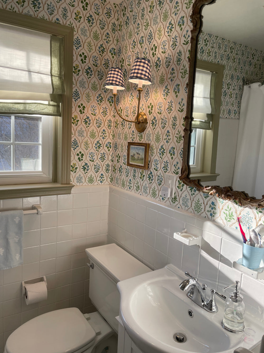 A vintage-style bathroom interior featuring a white pedestal sink, a white toilet, a wall-mounted double-light sconce with gingham lampshades, and wallpaper with a green and blue floral pattern on a cream background.