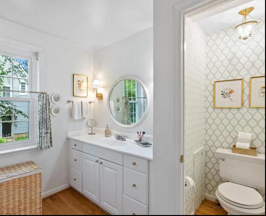 A bright white bathroom vanity with a round mirror, set in a room with light wood floors and a window. An open doorway on the right reveals an adjacent bathroom with green-and-white patterned tile, a toilet, and framed wall art.