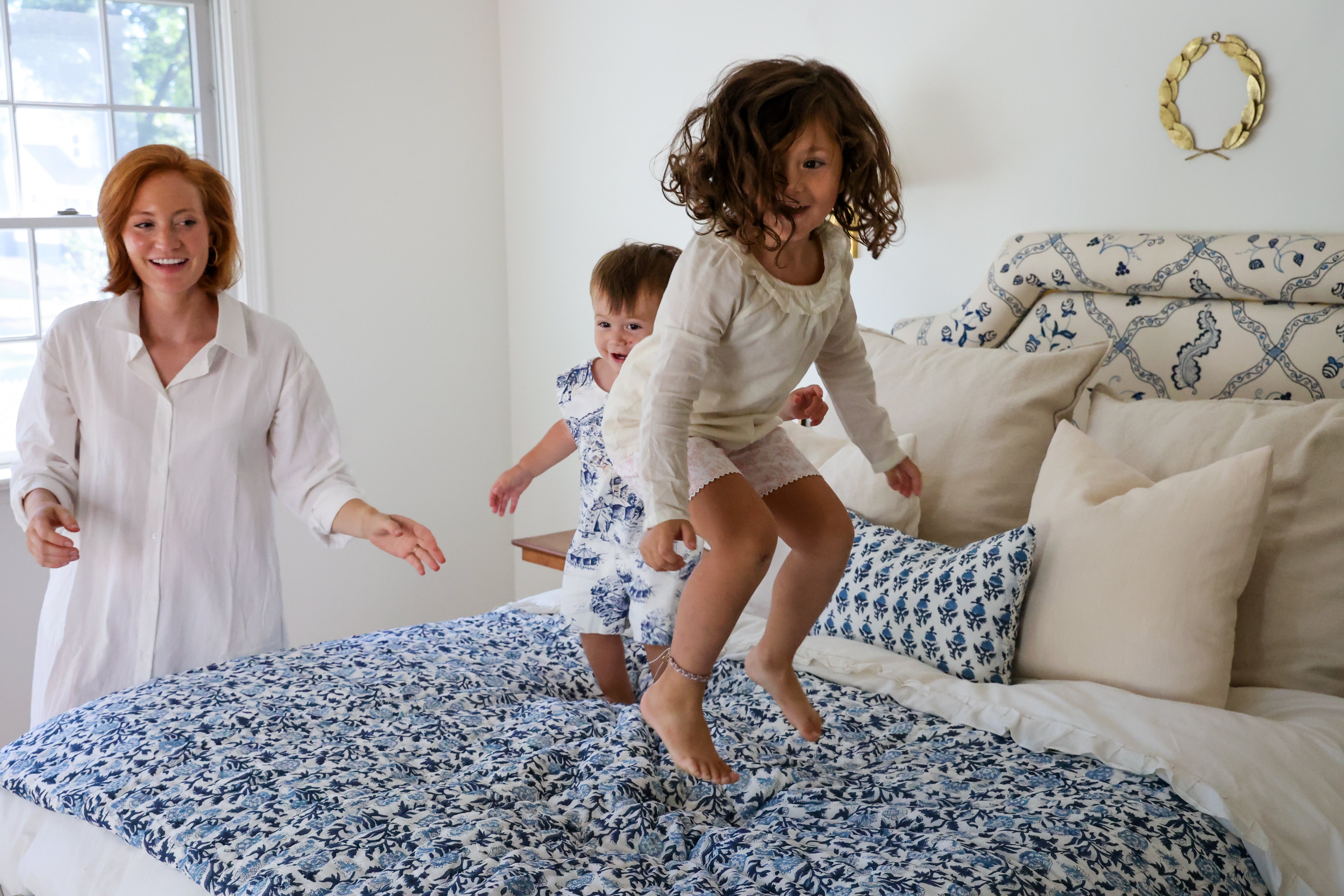 A woman and two young children are jumping on a bed with a blue and white patterned comforter and matching pillows in a bright bedroom.
