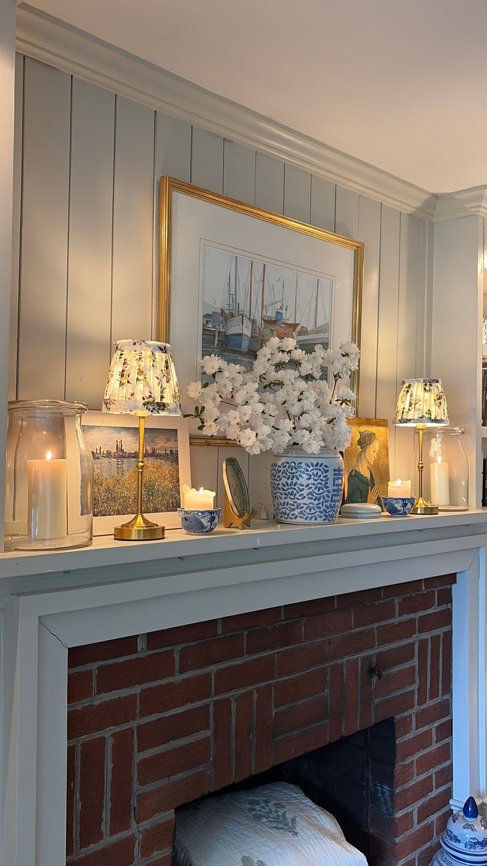 A cozy interior scene featuring a brick fireplace mantel decorated with a blue and white patterned vase of white flowers, small lamps, candles, and framed artwork against a white paneled wall.