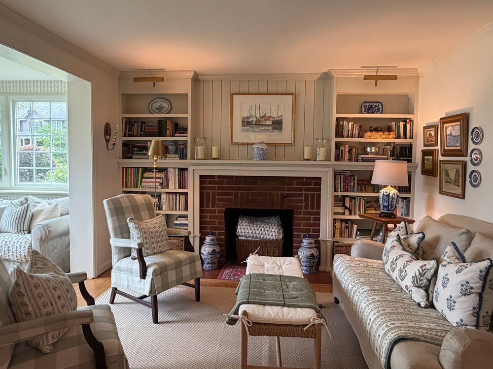 A cozy, traditional living room featuring a central brick fireplace with built-in bookshelves on either side, a beige sofa with patterned pillows, a plaid armchair, and a matching ottoman on a light rug.