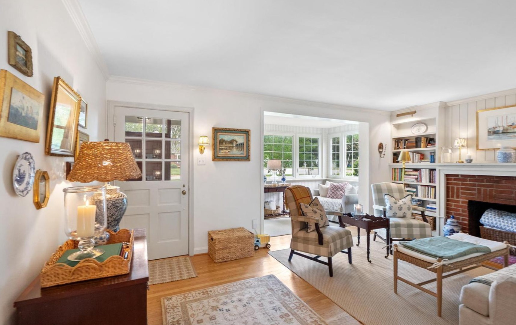 A bright, traditional living room with hardwood floors, a fireplace, a window seat, and multiple upholstered chairs and tables. The room is styled with framed art, lamps, and a patterned area rug.