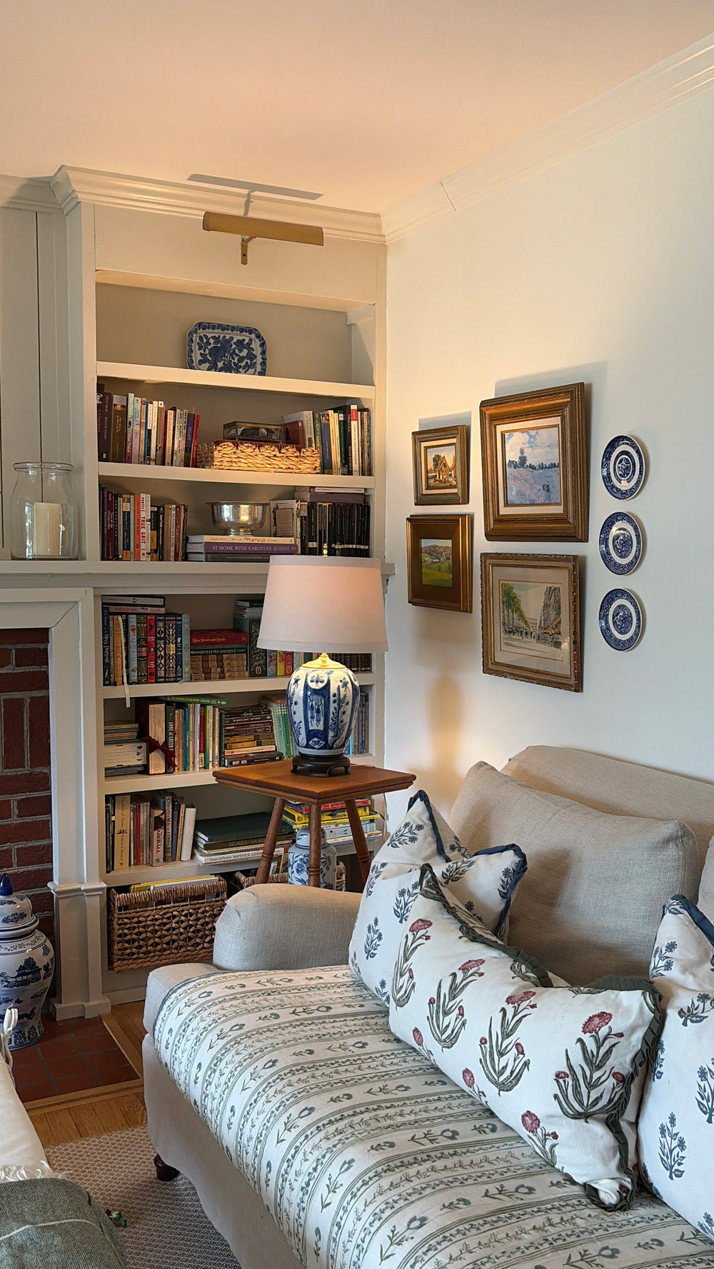 A cozy living room corner featuring a built-in bookshelf filled with books, a blue-and-white ceramic table lamp on a wooden side table, a beige sofa with patterned throw pillows, framed wall art, and a brick fireplace.