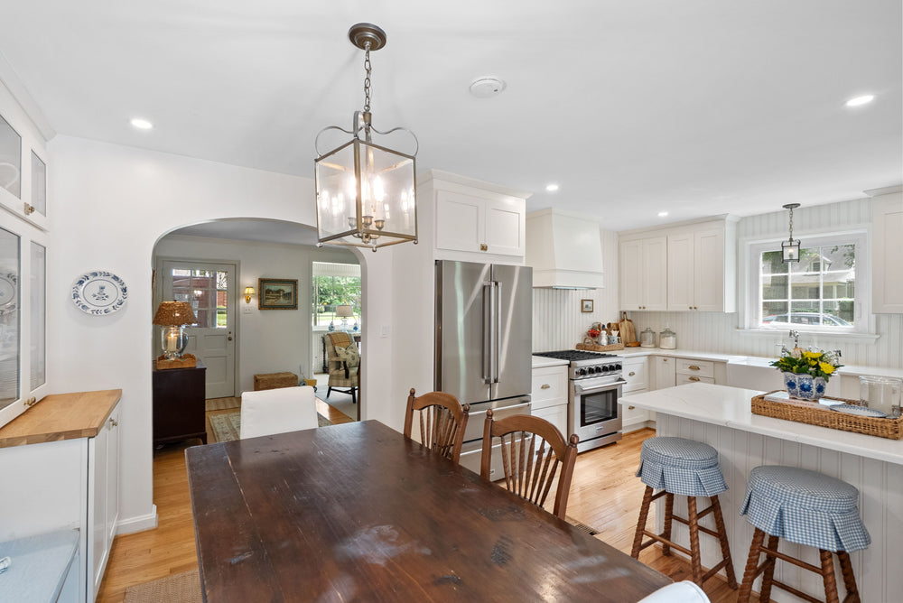 A bright, modern kitchen with white cabinets, a dark wood dining table, wooden bar stools with blue gingham cushions, stainless steel appliances, and a hanging lantern-style chandelier.