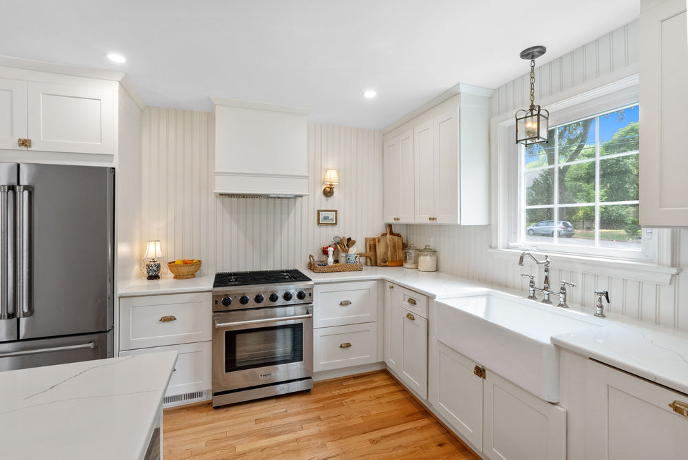 A bright, modern kitchen featuring white shaker-style cabinets, white countertops, a stainless steel gas range, a large white farmhouse sink under a window, and light wood floors.