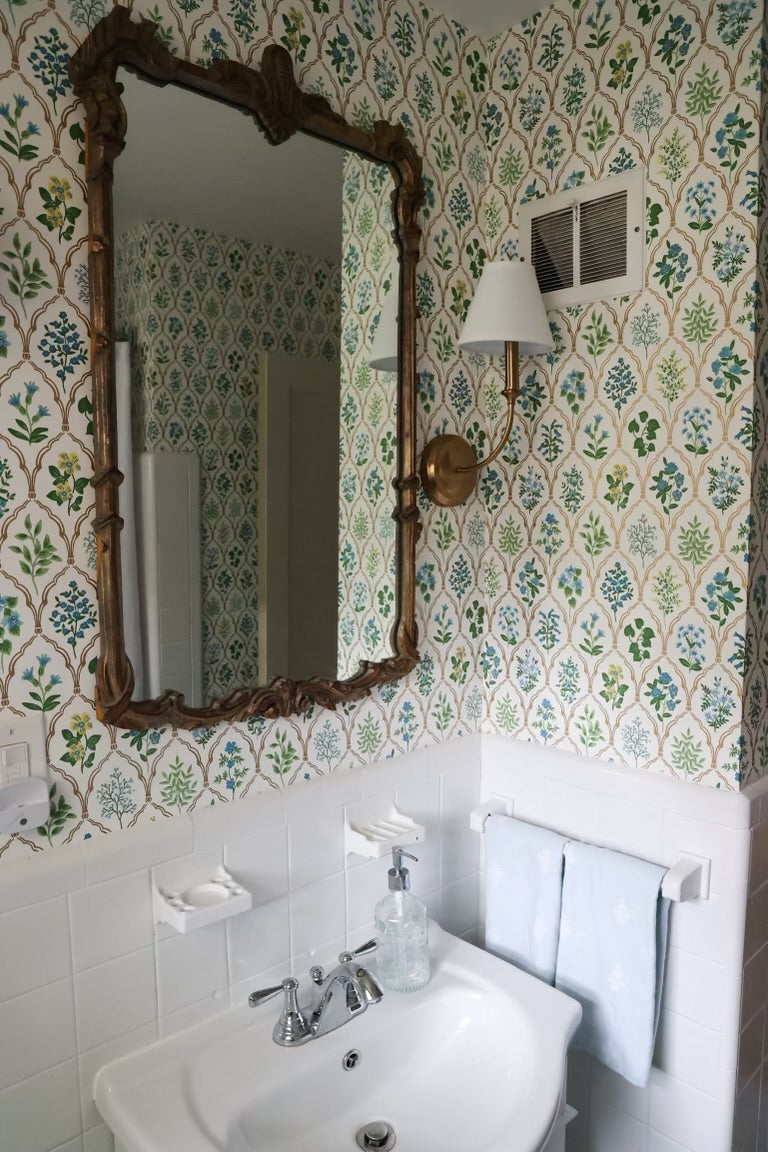 A vintage-style bathroom scene featuring a large ornate wooden mirror with a floral pattern wallpaper behind it, a white sink, and a wall-mounted brass sconce.