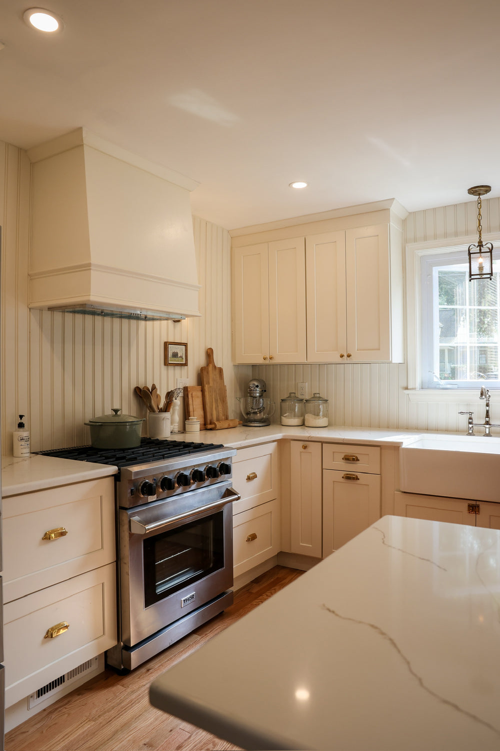 A bright, modern kitchen featuring cream shaker-style cabinets, a stainless steel gas range, a white farmhouse sink, and light wood flooring.
