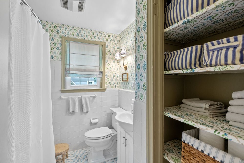 A bathroom interior with a white toilet, sink, mirror, shower curtain, patterned wallpaper, and built-in shelving on the right with striped and floral bedding and folded towels.