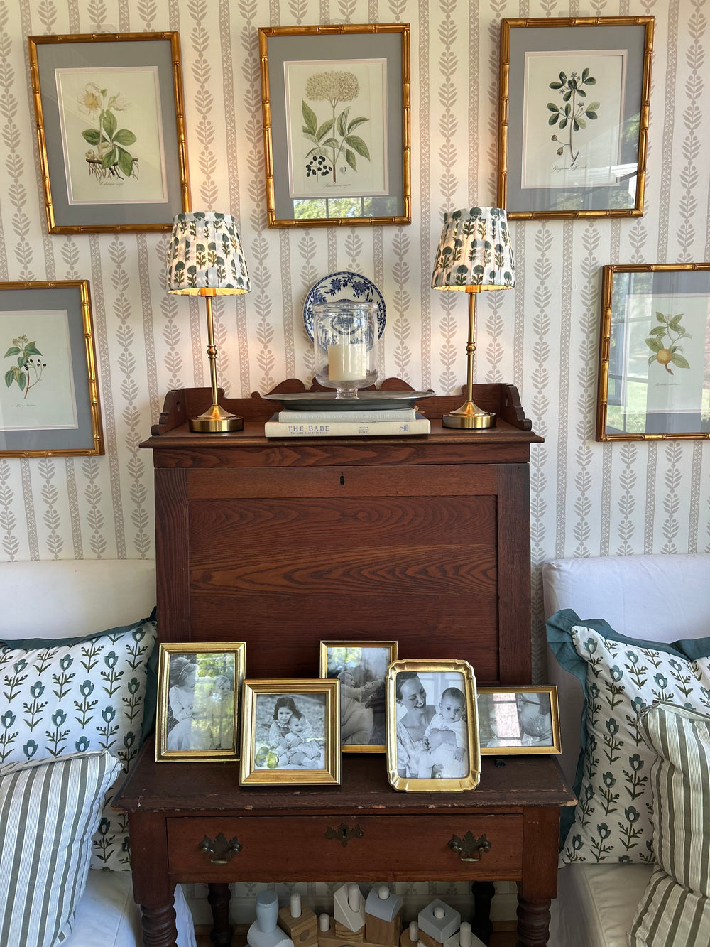 A dark brown wooden chest of drawers or cabinet with gold-tone hardware, styled in a room with botanical art prints on a patterned wallpaper wall, striped pillows, and matching lamps.